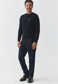 Marine kabelgebreide trui, donkerblauwe denim jeans, donkere schoenen. De trui heeft een klein geborduurd logo op de borst. Eenvoudige, klassieke pasvorm.
