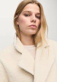 Abrigo de lana crema con una textura suave, solapas anchas y una silueta relajada. Debajo, se puede ver un top de punto ligero. Diseño sencillo y moderno.