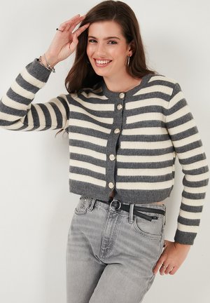Ung kvinde smilende, iført en grå og hvid stribet knap-up cardigan og lysegrå højhåndede jeans med sort bælte.