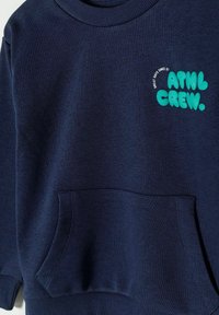 Tamsiai mėlyna sportinė palaidinė su banguotu apykakle, priekiu kišene ir žalia grafika, kurioje užrašyta "ATHL CREW" su tekstiniais akcentais.