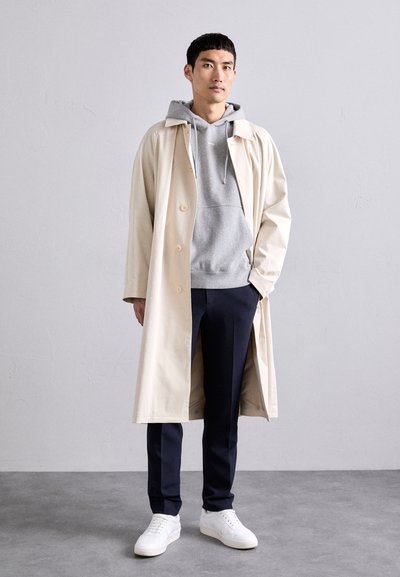 Manteau beige avec un design à col sur un sweat à capuche gris. Un pantalon sombre et des baskets blanches complètent le look. Silhouette simple et moderne.
