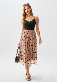 Schwarzes Rüschen-Camisole kombiniert mit einem nudefarbenen Tüllrock mit schwarzen Herzmustern. Das Modell hält eine schwarze Handtasche und trägt Sandalen mit Riemchen.