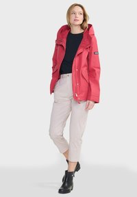 Rote wasserdichte Jacke mit Kapuze, ausgestattet mit Reißverschlusstaschen und einem Logo-Patch. Kombiniert mit hellen Hosen und schwarzen Knöchelstiefeln.