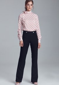 Kobieta stoi w różowej bluzce z długim rękawem w czerwone kropki oraz czarnych spodniacach z wysokim stanem na jednolitym szarym tle.