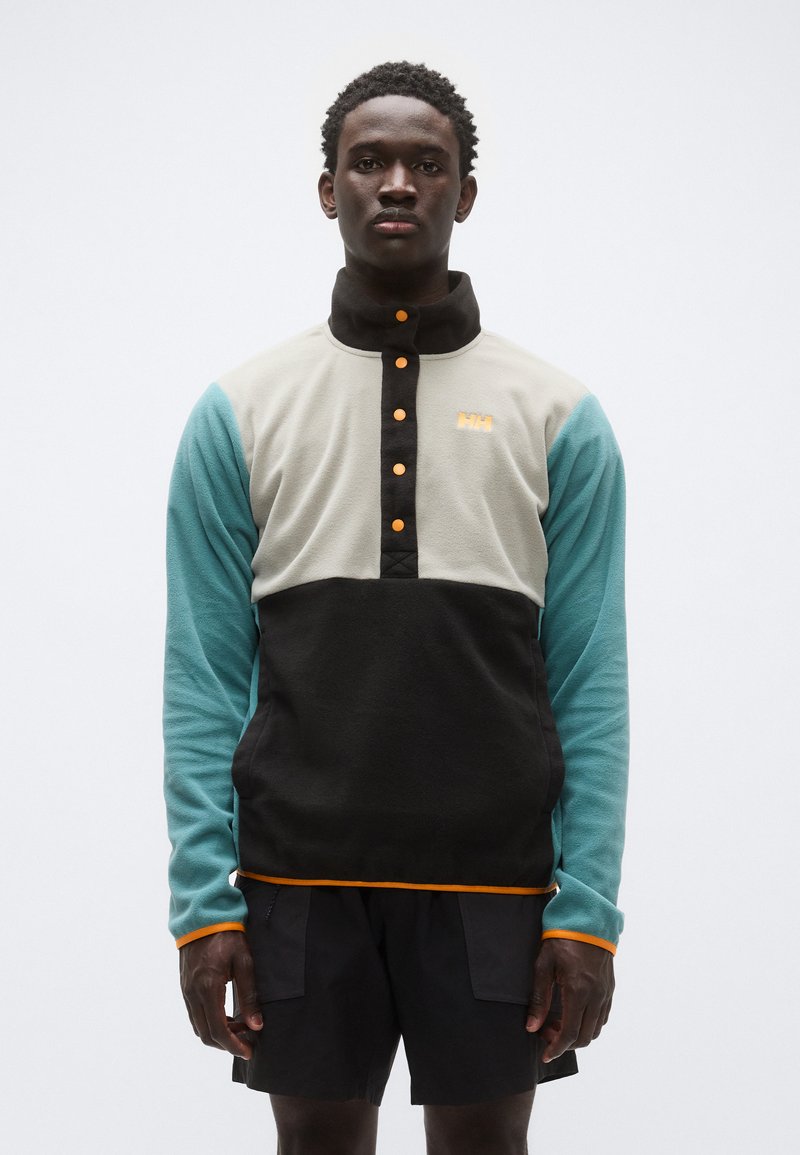 Φλις πουλόβερ με ψηλό γιακά, σε μπεζ, μαύρο και τιρκουάζ χρώματα, με πορτοκαλί λεπτομέρειες και κουμπιά πατ για το κλείσιμο μπροστά.