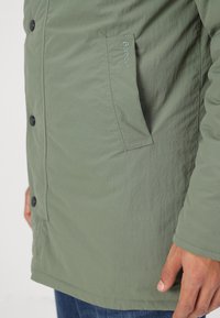 Olíva zöld, textúrált anyagú dzseki oldalsó zsebbel, gombos záródással és hímzett "Barbour" logóval a zsebén.