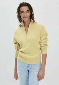 Gelber gestrickter Pullover mit hohem Kragen und silbernem Reißverschluss, mit gerippten Bündchen und Saum. Getragen über hellblauer Jeans.