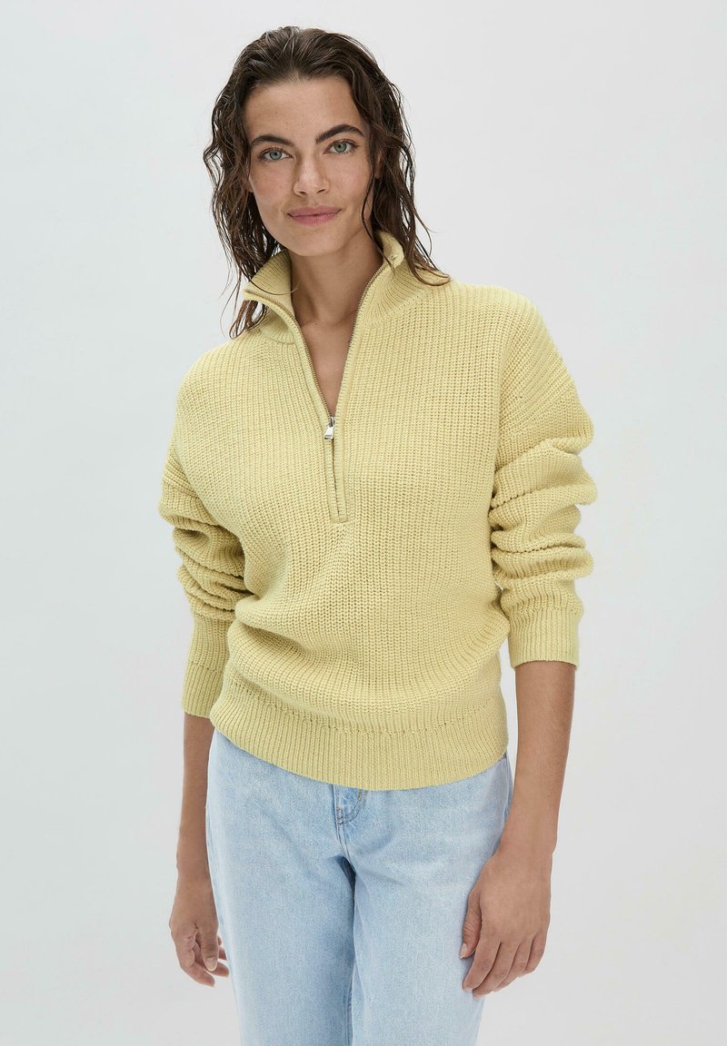 Gelber gestrickter Pullover mit hohem Kragen und silbernem Reißverschluss, mit gerippten Bündchen und Saum. Getragen über hellblauer Jeans.