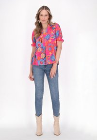 Blomstret bluse i livlig pink med korte puffærmer og v-udskæring. Sammen med tætsiddende blå jeans og beige cowboystøvler.