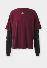 Bordeaux kortemouwshirt gelaagd over zwarte lange mouwen, met een textuurlogo op de achterkant en sterpatronen op de mouwen.