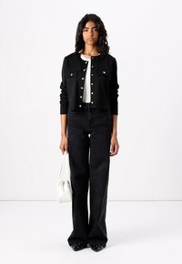 Schwarze, taillierte Jacke mit Knopfverschluss und zwei Vordertaschen. Darunter ein weißes, geripptes Shirt. Schwarze, ausgestellte Jeans und spitze Schuhe. Kleine weiße Tasche.