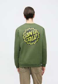 Άτομο με κοντά καστανά μαλλιά γυρισμένο πλάτη φορώντας φούτερ χακί με κίτρινο και μαύρο κυκλικό λογότυπο «Santa Cruz» στην πλάτη.