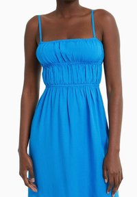 Žena v bezramenných jasně modrých šatech se skládaným živůtkem a tenkými ramínky, stojící s uvolněnými pažemi podél těla.