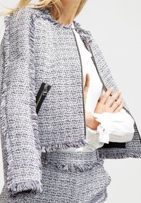 Kvinna klädd i en fransad tweedjacka och matchande shorts över en vit blus med volanger på ärmarna, poserar med armarna korsade.