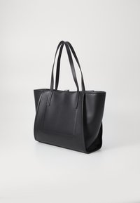 Sac fourre-tout en cuir noir avec deux poignées, forme rectangulaire, texture lisse et poche à patch à l'avant. Fermeture zippée en haut.