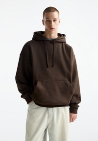 Brauner oversized Kapuzenpullover mit einer frontalen Tasche, Kordelzugkapuze und gerippten Bündchen, kombiniert mit hellen Hosen. Glatte Stoffoberfläche.