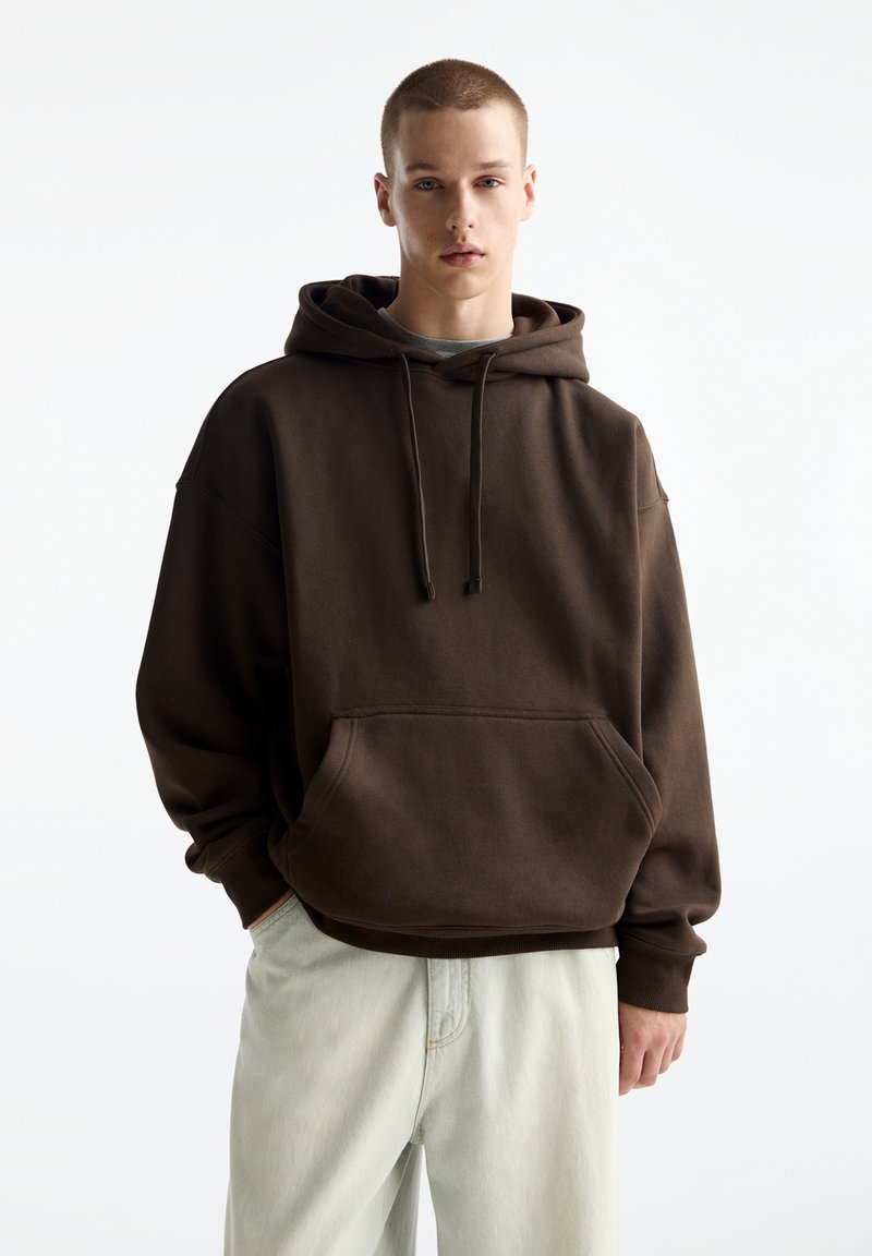 Brauner oversized Kapuzenpullover mit einer frontalen Tasche, Kordelzugkapuze und gerippten Bündchen, kombiniert mit hellen Hosen. Glatte Stoffoberfläche.