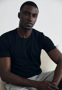 Schwarzes Baumwoll-T-Shirt mit kurzen Ärmeln, getragen von einer männlichen Figur. Er hat kurze Haare und sitzt vor einem hellen Hintergrund.