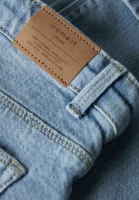 Lyseblå denimstoff med en brun lærlap med påskriften "name it DENIM" og synlige sømmer og beltesløyfe.