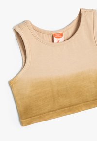 Vaalean beigeestä kultapruskeaan korostava tan-värinen puuvillatrikootoppi. Siinä on pyöreä pääntie ja hihaton malli. "Baby"-logo näkyy vastuuksena.