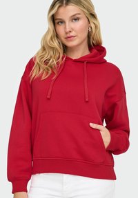 Femme aux longs cheveux blonds portant un sweatshirt à capuche rouge avec une poche avant et un pantalon blanc sur un fond uni.