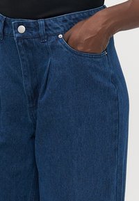 Temno modri jeans hlače z gumbom in zankami za pas, roka počiva v sprednjem desnem žepu, oblečene z črno majico.