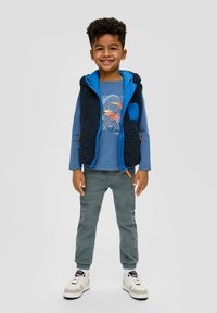 Kind trägt eine blaue Fleece-Weste über einem blauen Langarmshirt, grünen Hosen und weißen Sneakern mit schwarzen Akzenten. Lächelt.