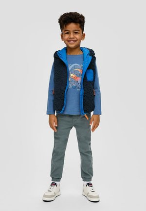 Kind dat een blauwe fleece-vest draagt over een blauwe long-sleeve shirt, groene broek en witte sneakers met zwarte accenten. Glimlachend.