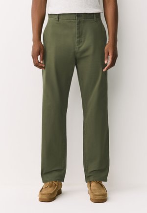 Férfi olívazöld, egyenes szárú nadrágot visel gombos rögzítéssel és világos barna velúr fűzős cipőket, egy egyszerű háttér előtt áll.