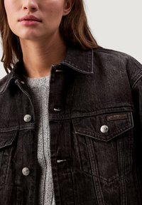 Černá džínová bunda s knoflíkovým zapínáním, náprsními kapsami s klopou a štítkem Calvin Klein, nošená přes světle šedý texturovaný pletený svetr.