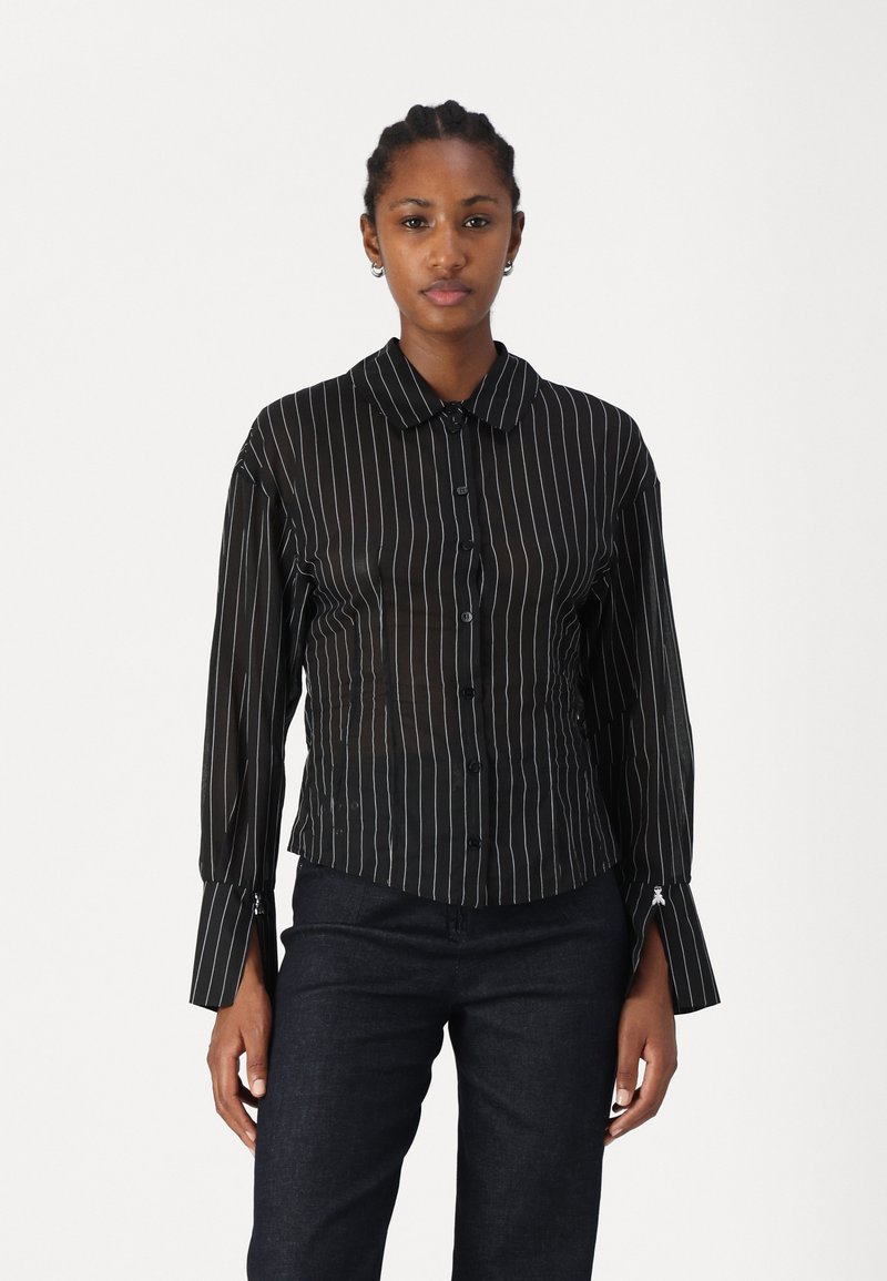 Camicia nera con bottoni, a righe verticali bianche, maniche lunghe e colletto. Caratterizzata da un design aderente e polsini con dettagli unici.