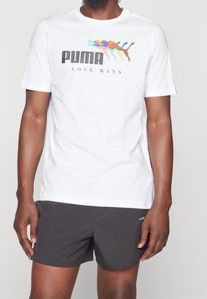 Mand iført hvid Puma T-shirt med farverigt springende puma-logo og teksten "Love Wins", kombineret med sorte Puma-shorts.