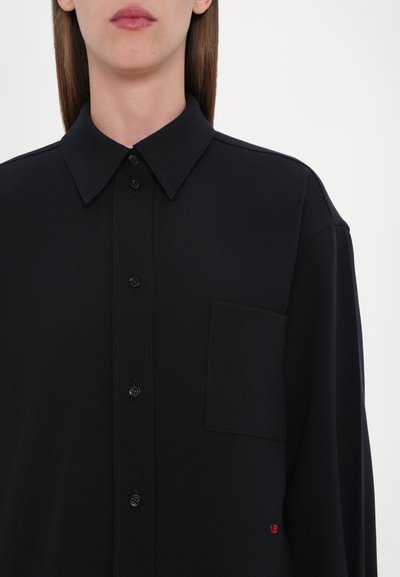 Camicia nera con bottone, dotata di colletto strutturato, taschino frontale e tessuto leggermente testurizzato. Presenta bottoni neri e un piccolo dettaglio con logo rosso.