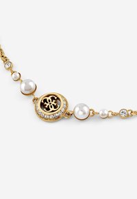 Goudkleurige ketting met een cirkelvormige hanger met een logo, omringd door witte parels en kleine heldere kristallen, aan een schakelkettinkje.