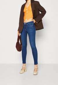 Jeans ajustados azules, top de encaje naranja, blazer oversized marrón y zapatos de tacón bloque crema. Lleva un bolso texturizado marrón.