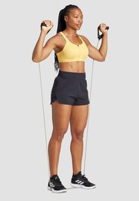 Dzeltena sporta krūšturis ar priekšējo aizdari, melni sporta šorti ar tuvējo jostas daļu un melnas čības ar baltajiem akcentiem, turēdamas pretestības joslas.