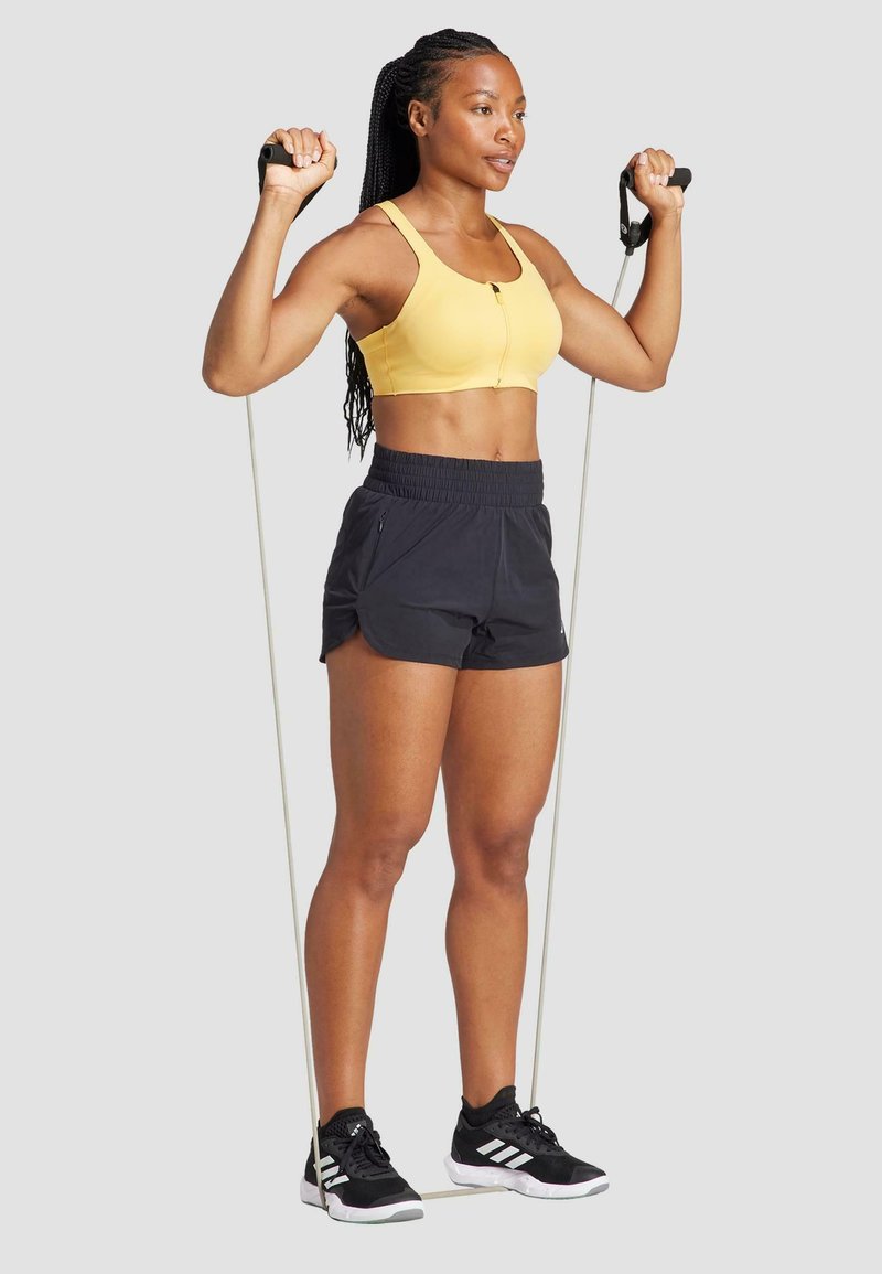 Dzeltena sporta krūšturis ar priekšējo aizdari, melni sporta šorti ar tuvējo jostas daļu un melnas čības ar baltajiem akcentiem, turēdamas pretestības joslas.