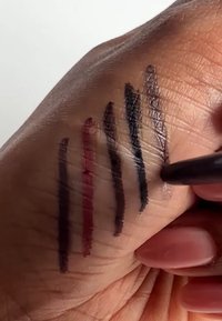 Proben von sechs Eyeliner-Farben auf einer Hand aufgetragen, darunter Rot, Dunkellila und Schwarz, mit glatten Texturen und unterschiedlichen Breiten.