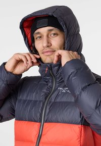Padded Jacke in marineblau und leuchtend rot, mit hohem Kragen, durchgehendem Reißverschluss und abnehmbarer Kapuze mit kontrastierender Innenausstattung.