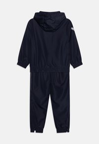 Survêtement bleu marine en tissu lisse et léger, doté d'une capuche, de poignets élastiques et d'ouvertures de jambe zippées.