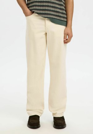 Personne portant un pantalon ample couleur crème, des chaussures marron foncé et un haut en maille rayé avec une main dans la poche sur fond blanc.