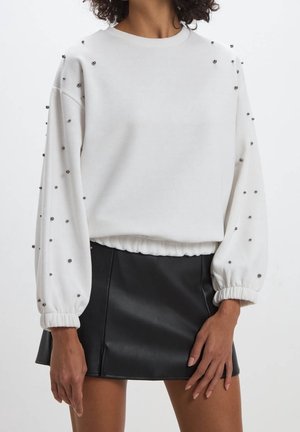Vrouw draagt een wit sweatshirt met zilveren studs op de mouwen en een zwarte leren minirok tegen een effen achtergrond.