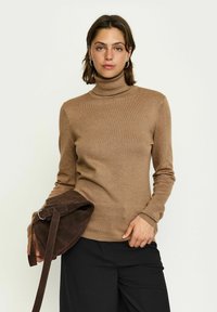 Maglione a collo alto marrone in tessuto a coste, con maniche lunghe. Il modello tiene in mano una borsa in suede marrone scuro con tracolla. Sfondo semplice.