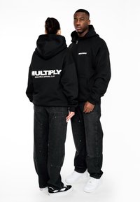 Černé mikiny s nápisem "MULTIPLY" a "MULTIPLY APPAREL CLUB" v bílé barvě. Nazuté s tmavými džíny a teniskami, ukazující uvolněný střih.