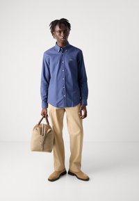 Marineblauwe overhemd met knopen en een logo, beige broek, bruine suède schoenen en een tan kleurige weekendtas met rits en bruine accenten.