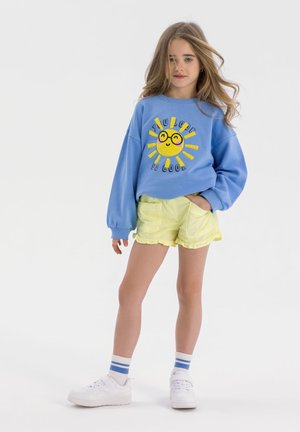 Sweatshirt bleu avec graphique de soleil et texte, associé à un short jaune avec un ourlet à volants. Baskets blanches et chaussettes rayées complètent le look.