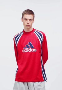 Maillot de sport rouge en mesh à manches longues avec des rayures latérales bleu marine et blanches, arborant un grand logo Adidas central sur le devant.