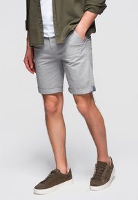 Homme portant un short gris clair retroussé, une chemise vert olive à manches retroussées, un t-shirt blanc et des baskets marron, debout devant un fond uni.