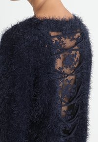 Marineblauer, flauschiger Pullover mit einem Spitzenrücken, der florale Muster und gekreuzte Details zeigt. Weiche Textur und Rückenausschnitt-Design.