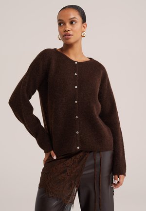 Femme portant un pull boutonné marron foncé, des boucles d'oreilles créoles dorées et un pantalon foncé avec un détail en dentelle marron sur un côté.